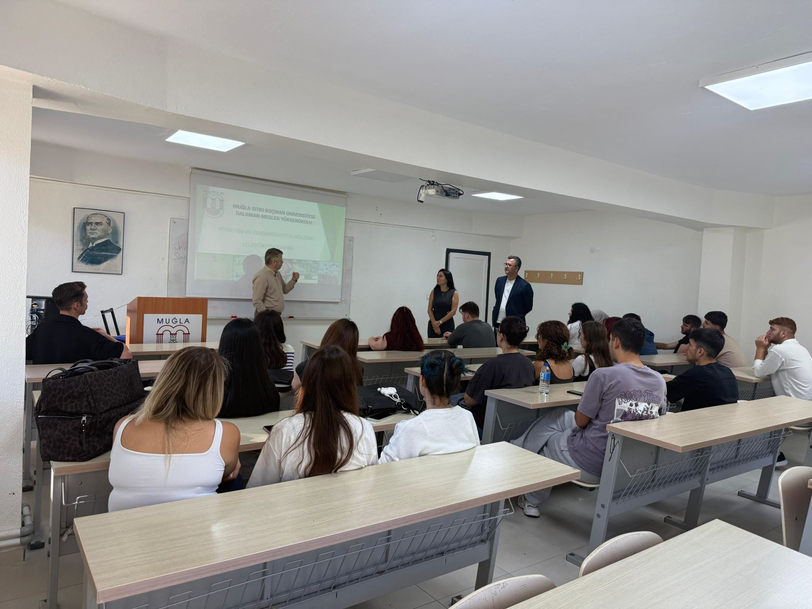 Derslikte açılış-tanıtım sunumu; projeksiyonda program başlığı, bir konuşmacı sahnede, iki kişi kenarda, öğrenciler sıralarda.